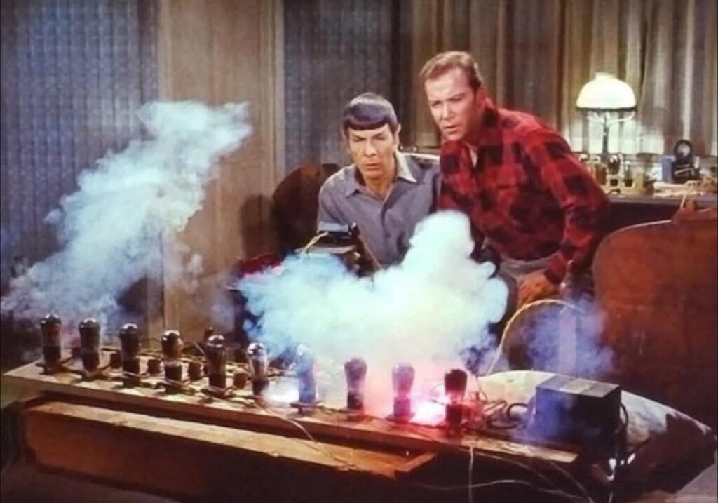 Spock and Capt. Kirk are leaning over a wooden platform filled with electronic components. Smoke is billowing from the setup, creating a mysterious and intense atmosphere. They appear to be concentrating on the experiment.