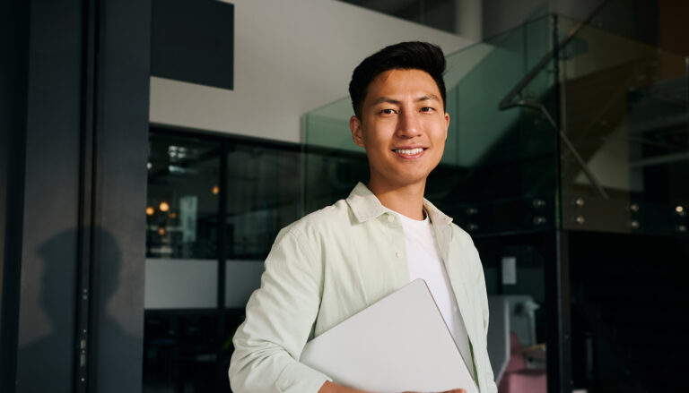 A smiling man in a light green shirt holds a laptop while standing in a modern office with glass walls and a staircase in the background.